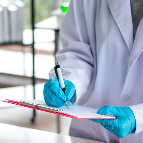 Person wearing a lab coat and gloves writing on a clipboard.